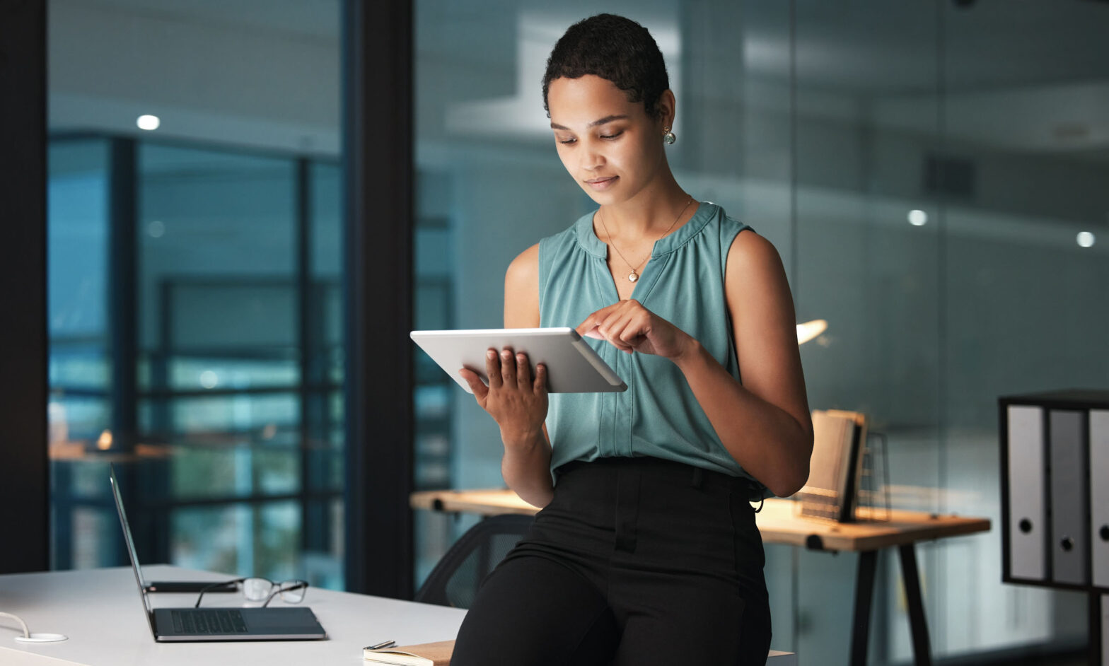 A woman dressed professionally is sat on the edge of a desk, looking at a digital tablet.